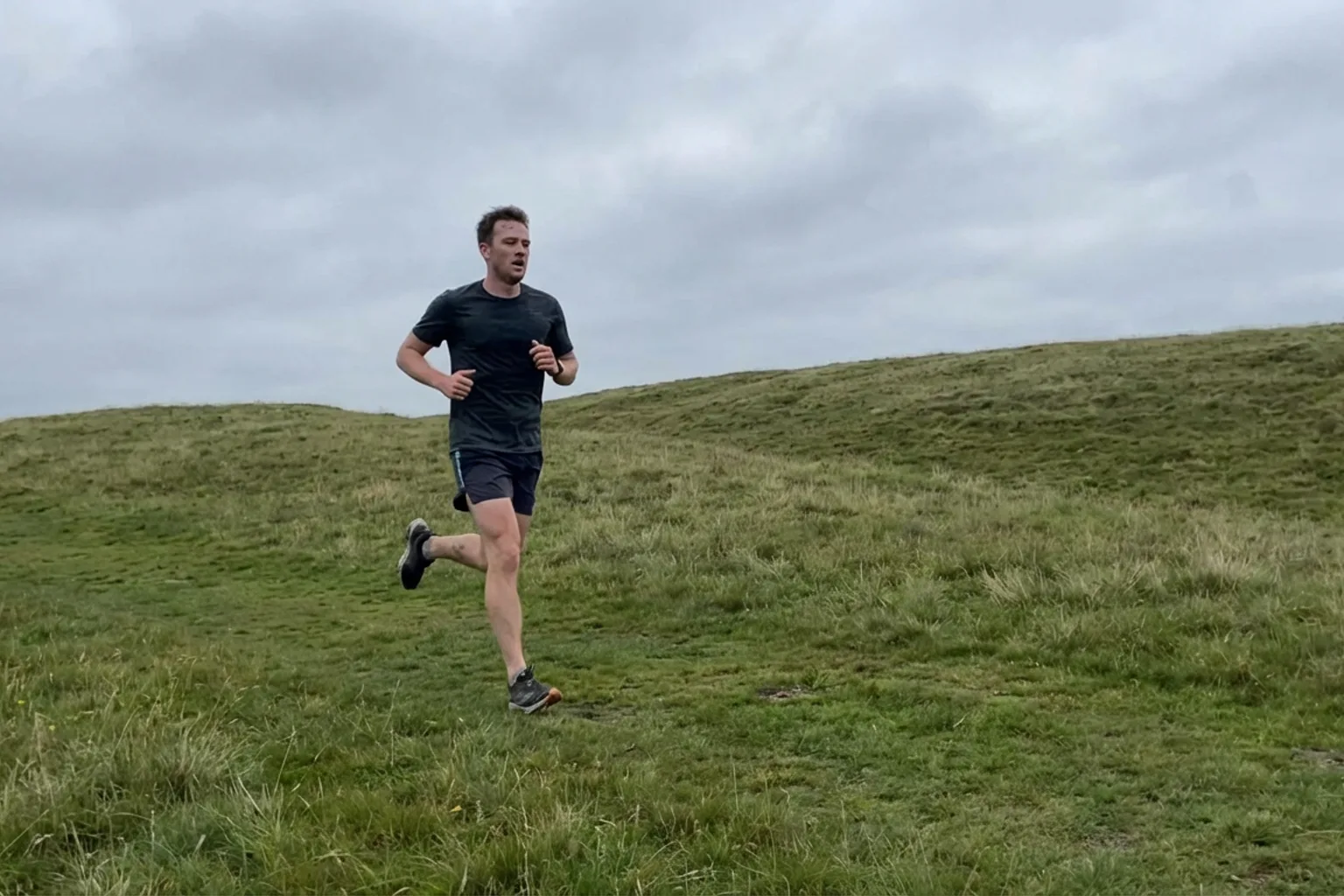 Candid iPhone photo of a fit male runner on a gentle grass hill, mid-stride going uphill, natural overcast light, casual