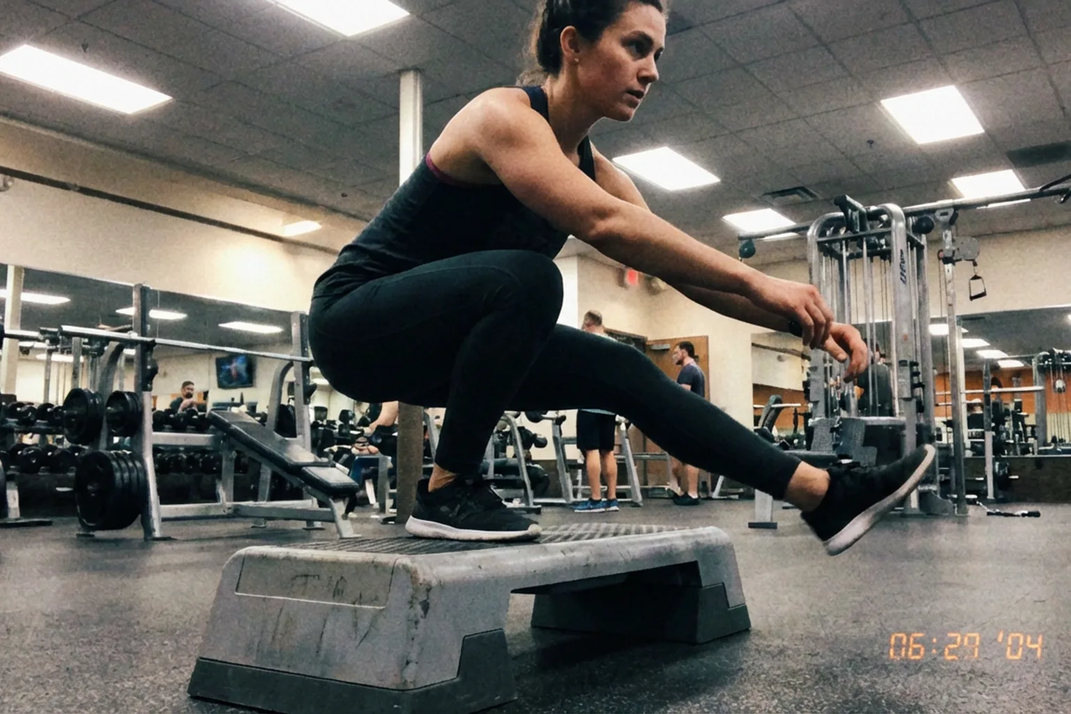 Candid iPhone photo of an athletic woman doing a single-leg squat exercise on a gym step, natural overhead lighting, rea