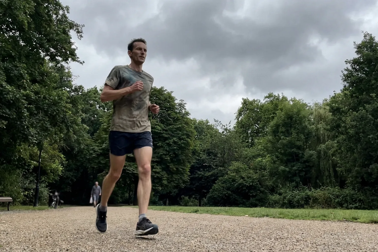 Candid iPhone photo of a lean male runner mid-stride on a tree-lined park path, slightly off-centre framing, overcast na