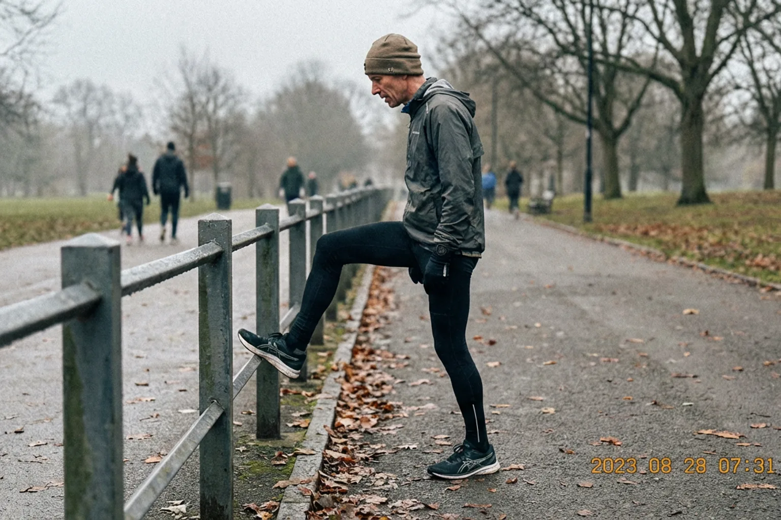 Candid iPhone photo of a lean male runner in his mid-40s stretching his calf against a park railing, overcast morning li