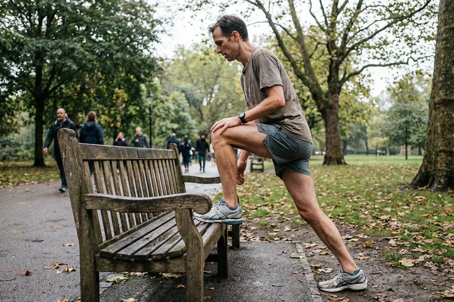 Candid iPhone photo of a lean male runner stretching his hip and glute against a park bench, natural light, overcast sky